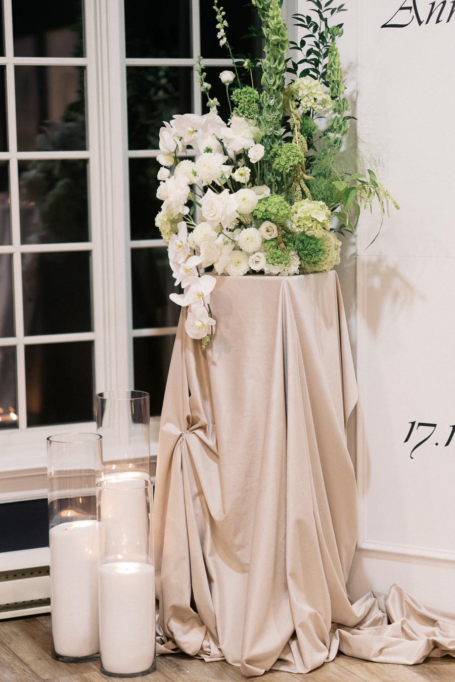 Elegant floral arrangement with white and green blossoms atop a draped beige fabric, accompanied by tall glass candles, situated in front of window panes and a white wall.