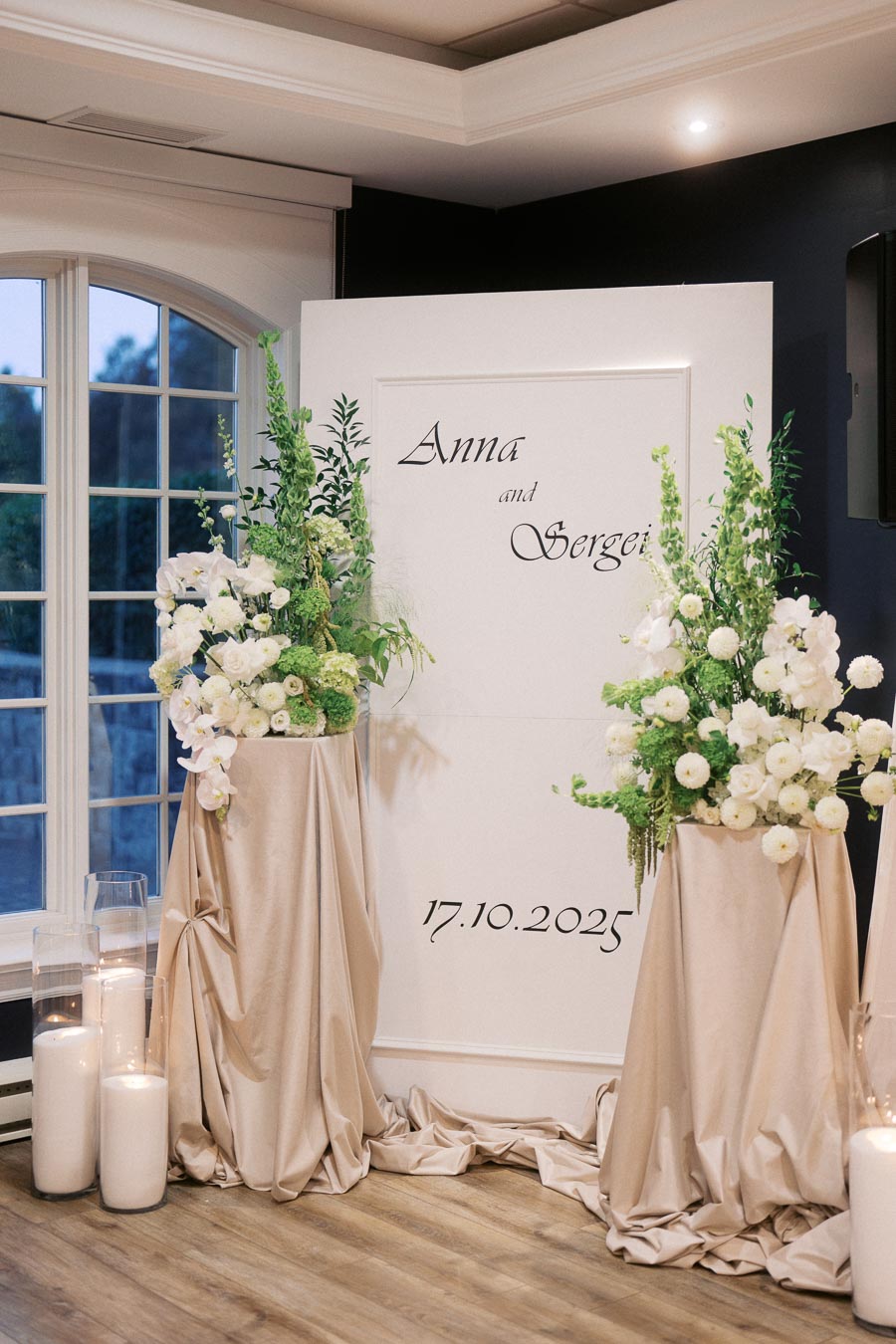 Elegant wedding decor featuring a personalized backdrop with the names Anna and Sergei and the date 17.10.2025, adorned with lush white and green floral arrangements and soft candle lighting, set in a sophisticated indoor venue space.