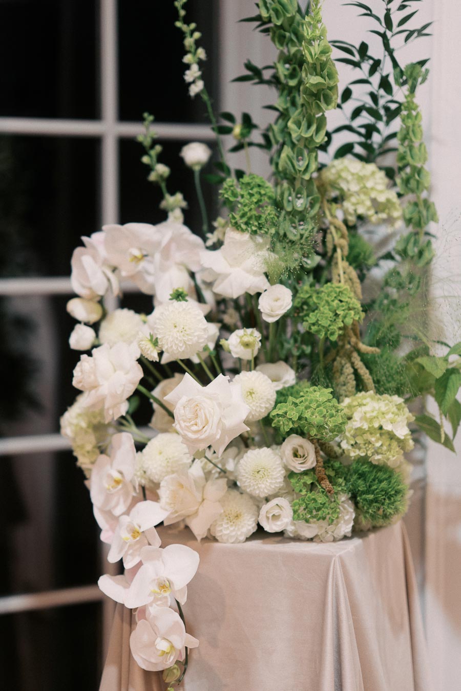 Elegant floral arrangement with white roses, hydrangeas, and lush greenery atop a draped table, semi-formal setting.
