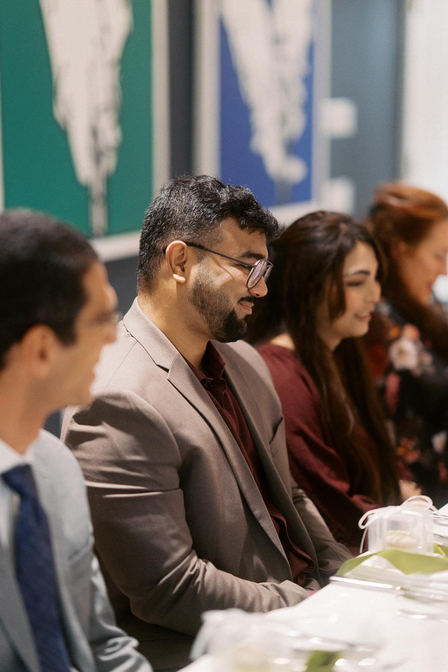 Group of people sitting at a formal meeting, dressed in business attire, with focus on a man wearing glasses and a suit, smiling and engaging in conversation. Blurred background with colorful artwork on the wall.