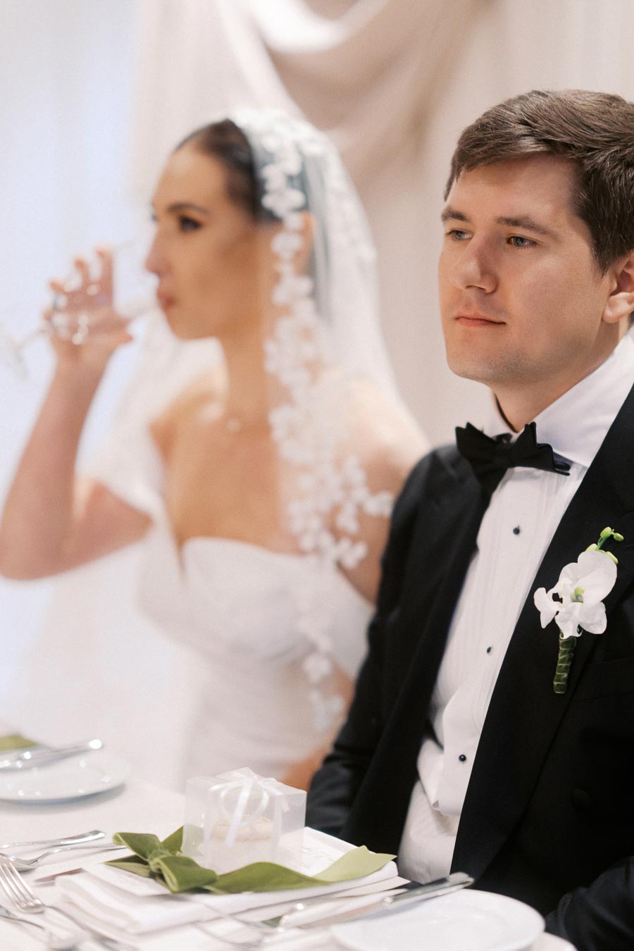Bride and groom seated at wedding reception table, groom wearing black tuxedo with white boutonniere, bride in white dress with floral veil, drinking from a glass, elegant table setting with gift box and green napkin.