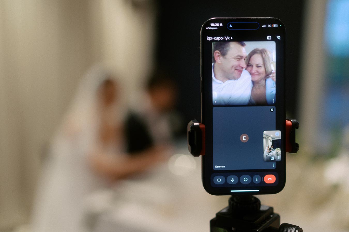 Smartphone on a tripod showing a video call with a couple smiling, blurred background of a bride and groom at a wedding.