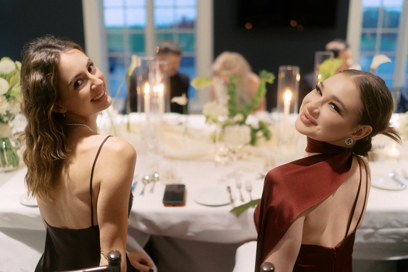 Two smiling women in elegant evening dresses seated at a beautifully set dinner table with candles and floral arrangements, creating a warm and sophisticated ambiance.