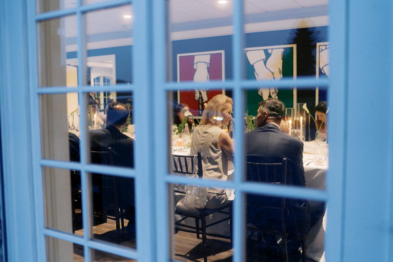 View through a window of a formal dinner gathering with elegantly dressed guests seated at a candlelit table, featuring modern artwork on blue walls in the background.