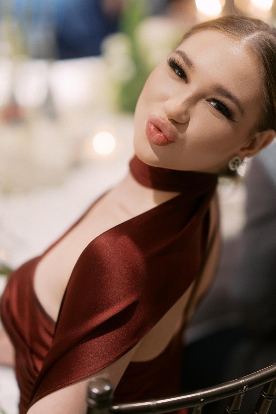 Young woman in an elegant burgundy dress, sitting at a formal event, playfully purses lips for the camera.