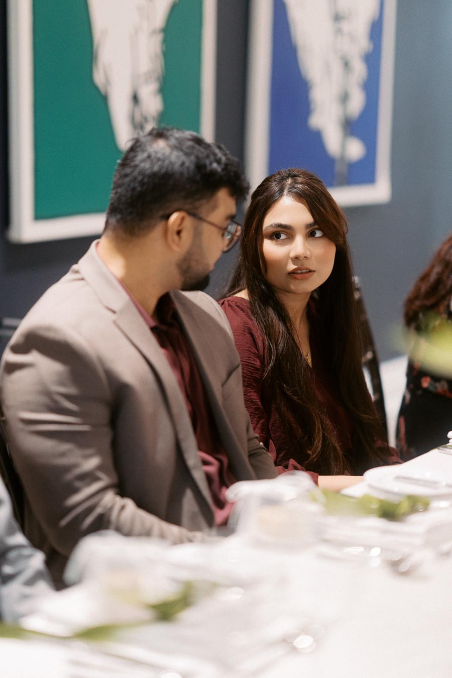 A man and a woman engaged in a conversation during a formal dinner setting, featuring elegant tableware and modern art in the background.