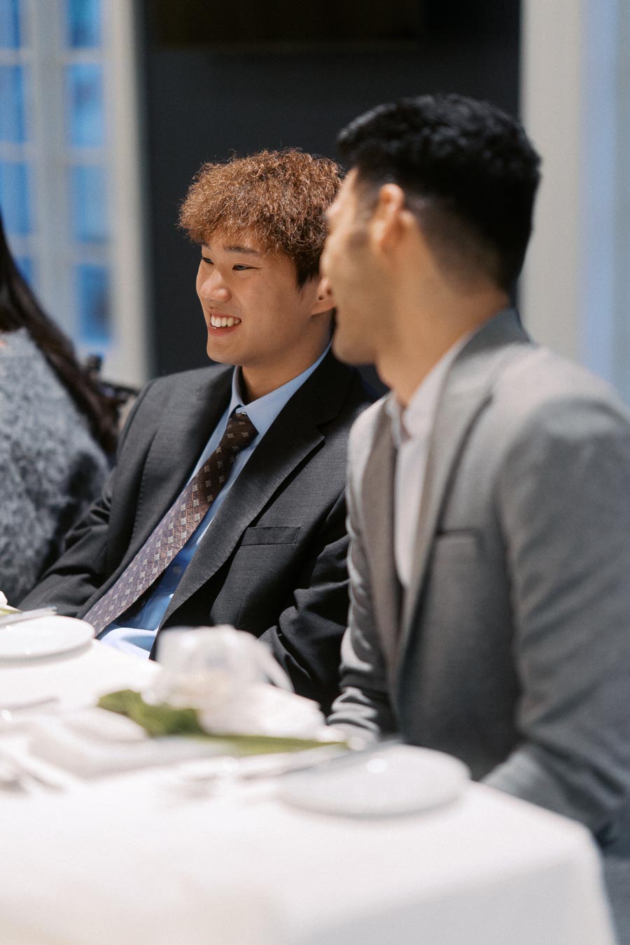 Two people in formal attire smiling and conversing at a dining table set with white linens and dishes, in a well-lit indoor setting.