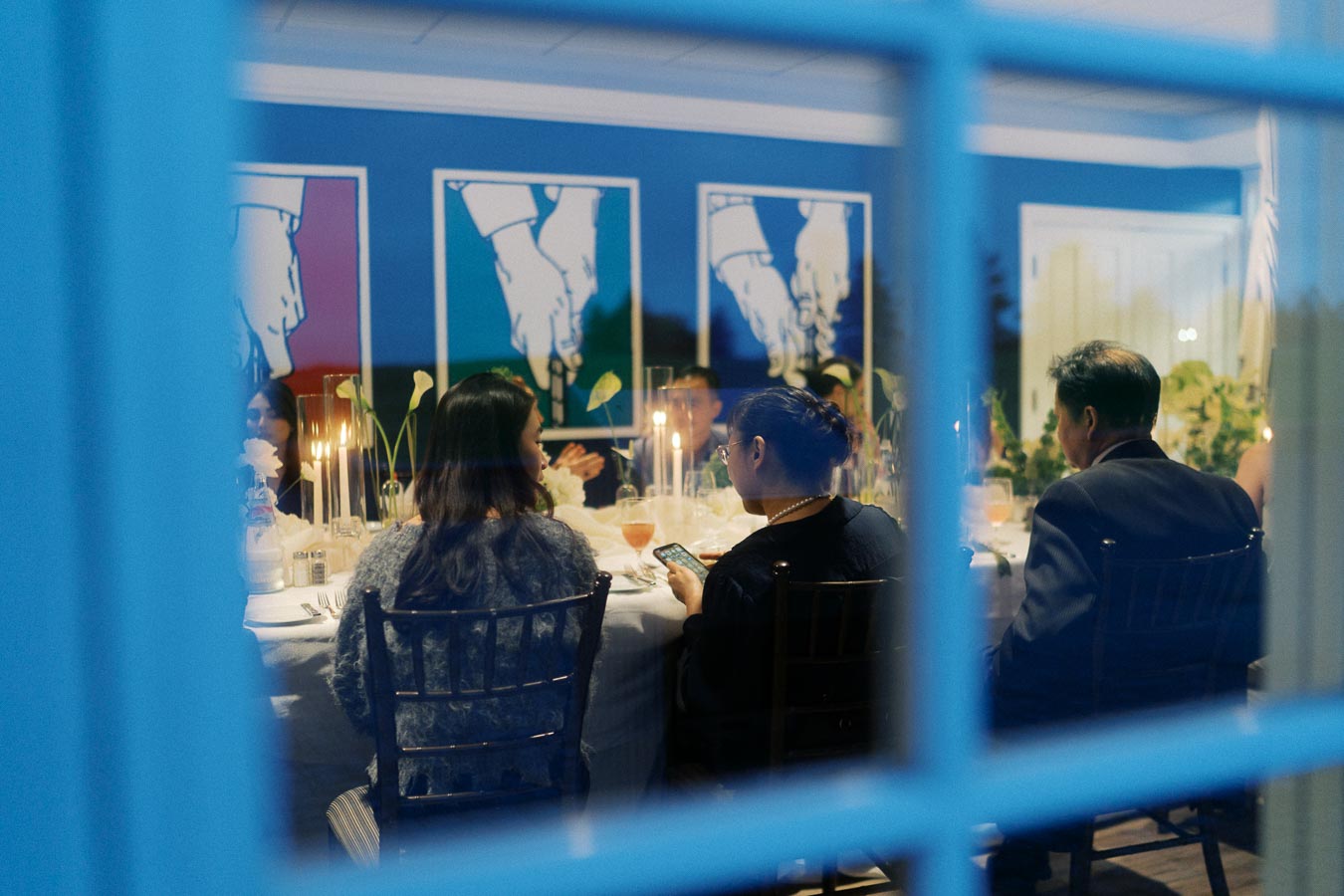 View through a blue-framed window of a formal dinner gathering. Guests seated around a table engage with each other, with elegant table settings and floral centerpieces visible. Wall art featuring abstract hand illustrations adds a modern touch to the sophisticated dining atmosphere.