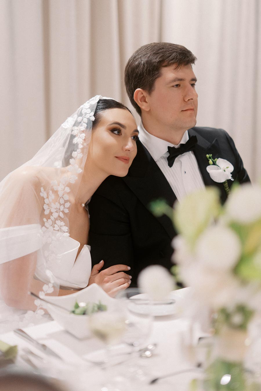 Bride and groom sitting together at a wedding reception, with the bride wearing a floral lace veil and the groom in a black tuxedo.