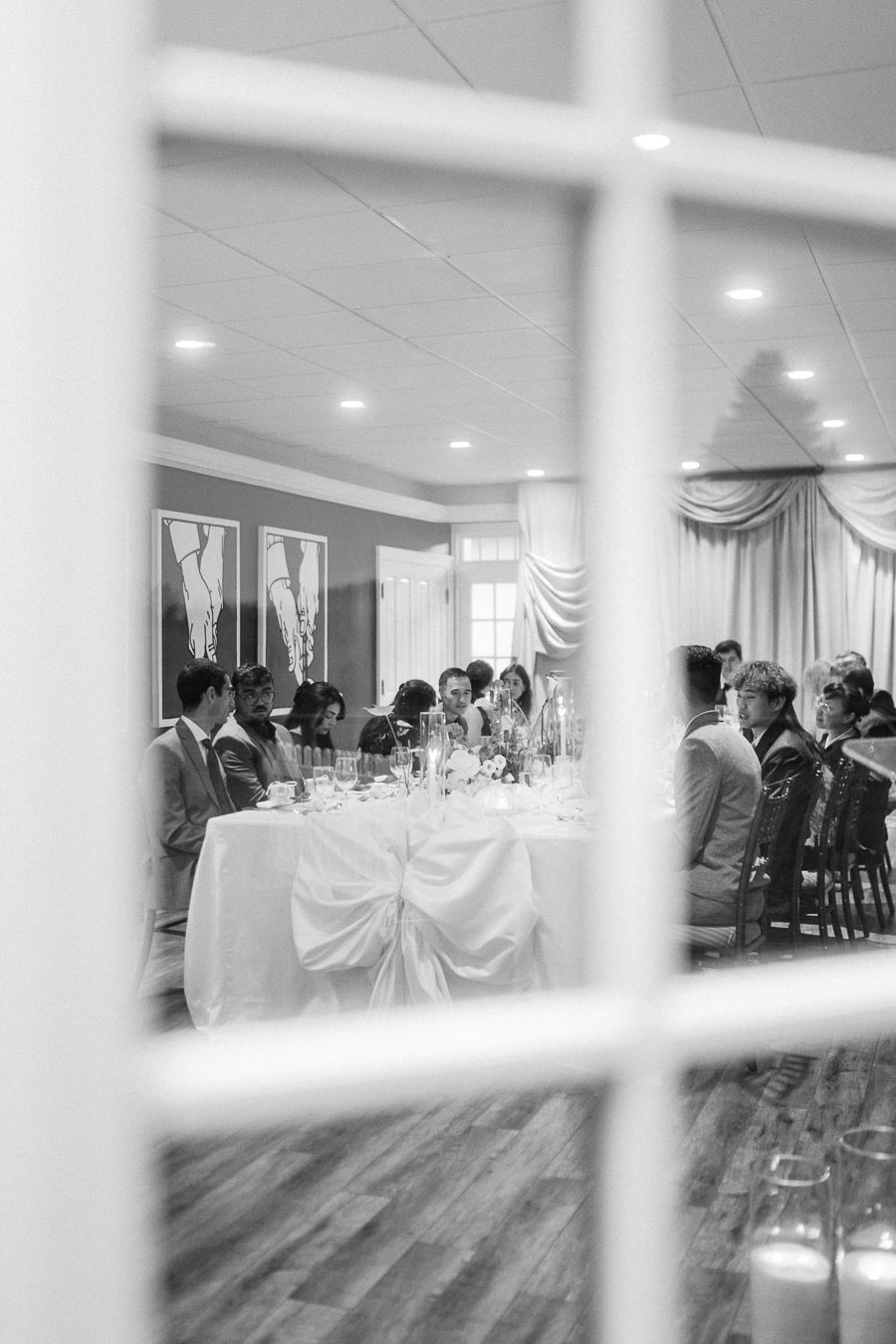 Black and white photo of a formal dining event viewed through a windowpane, featuring elegantly dressed guests seated around a decorated table with candles and floral centerpieces.