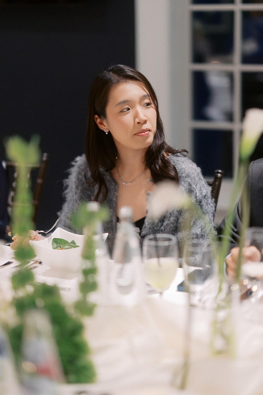 Young woman sitting at a dining table, holding a fork with a salad dish in front of her. The scene includes elegant tableware and glasses, suggesting a formal dining setting.