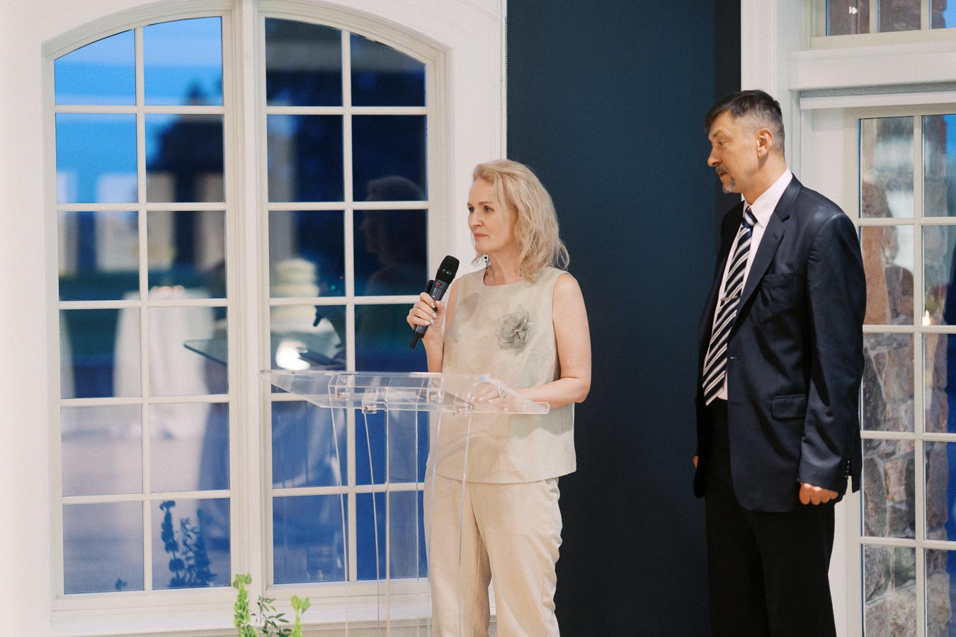 A woman speaking at a clear podium with a microphone, dressed in light attire, with a man in a dark suit standing beside her, in front of large windows indoors.