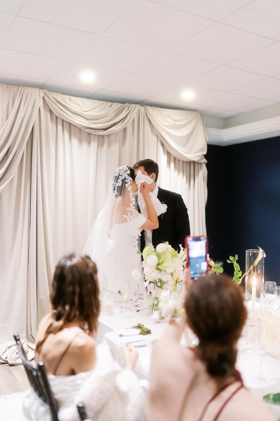 Bride and groom sharing a kiss at their wedding reception, with guests capturing the moment on a smartphone. Elegant white drapery and floral table centerpiece enhance the romantic atmosphere.