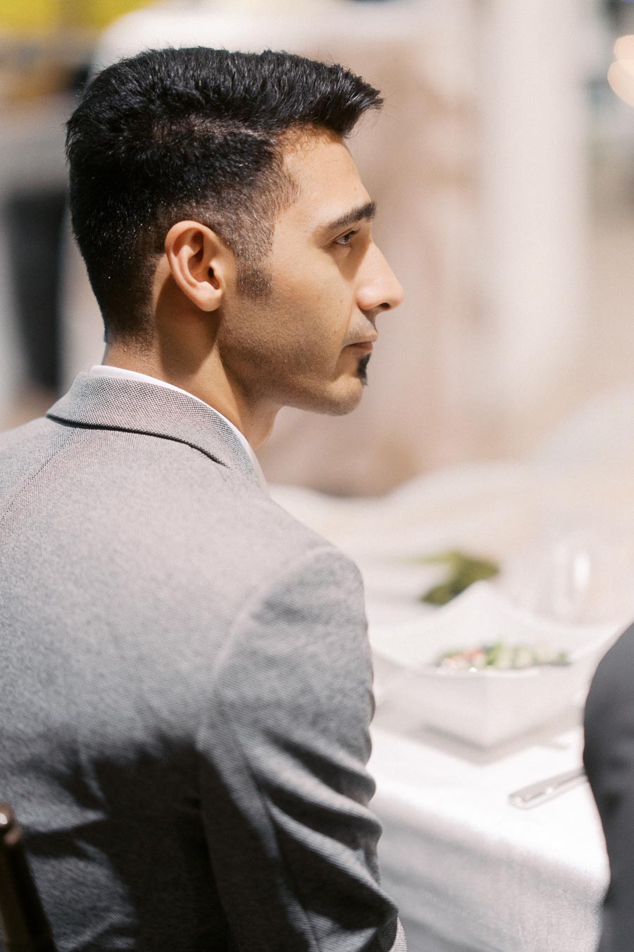 Profile view of a well-dressed man seated at a table, wearing a grey suit, with a blurred background and a focus on his serious expression.