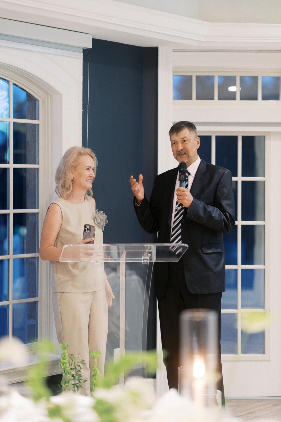 A man and a woman stand at a clear podium during a formal event. The man is speaking into a microphone, and the woman holds a phone, smiling warmly. They are inside a well-lit room with large windows and elegant decor.
