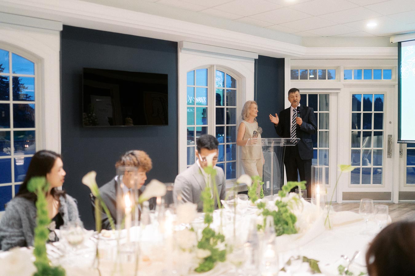 A formal event with elegantly dressed guests seated at a long dining table decorated with white flowers and candles; a man and woman stand at a podium delivering a speech in a well-lit room with large windows.