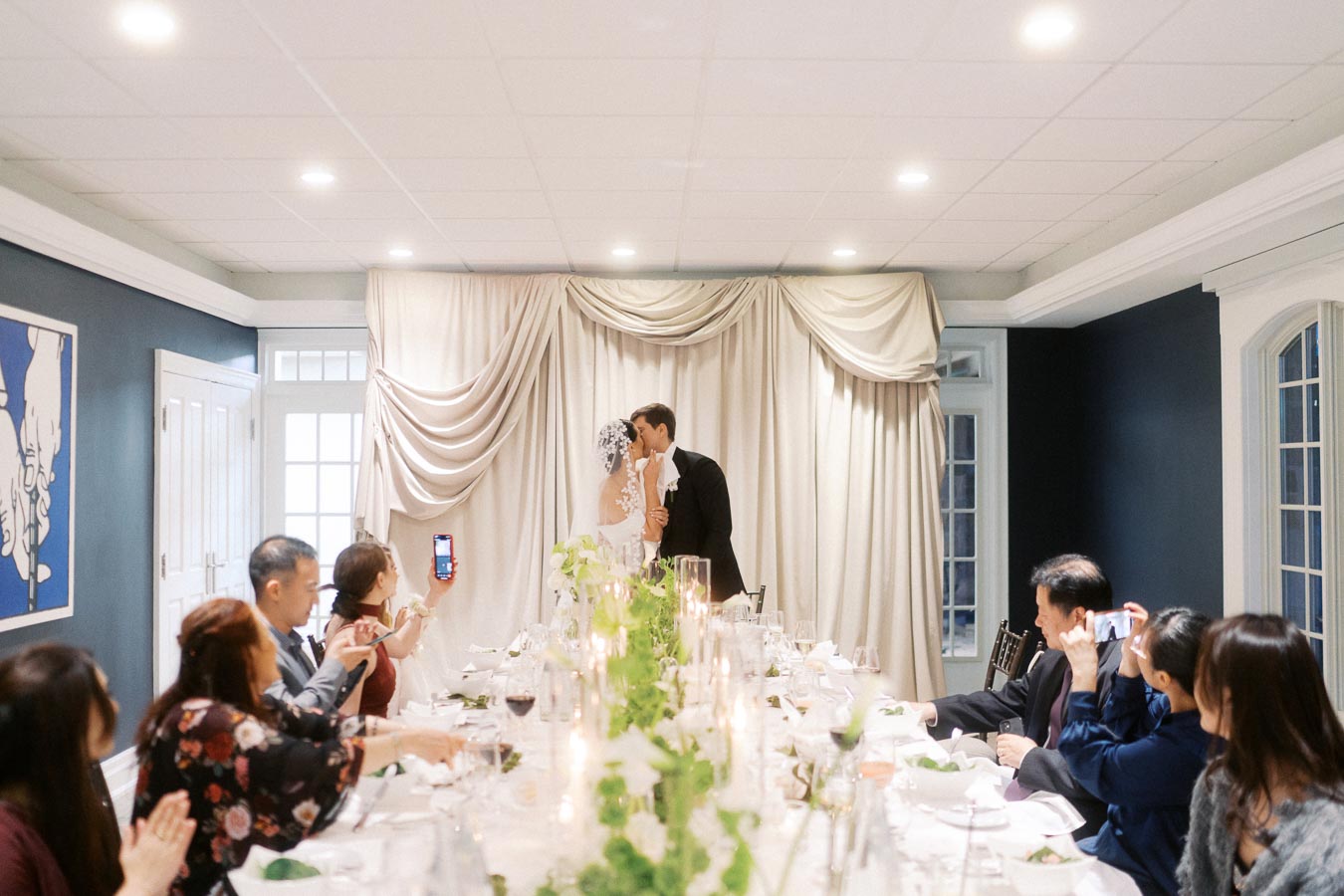 Bride and groom sharing a kiss at the head of an elegantly decorated wedding reception table, surrounded by guests taking photos and enjoying the celebration.