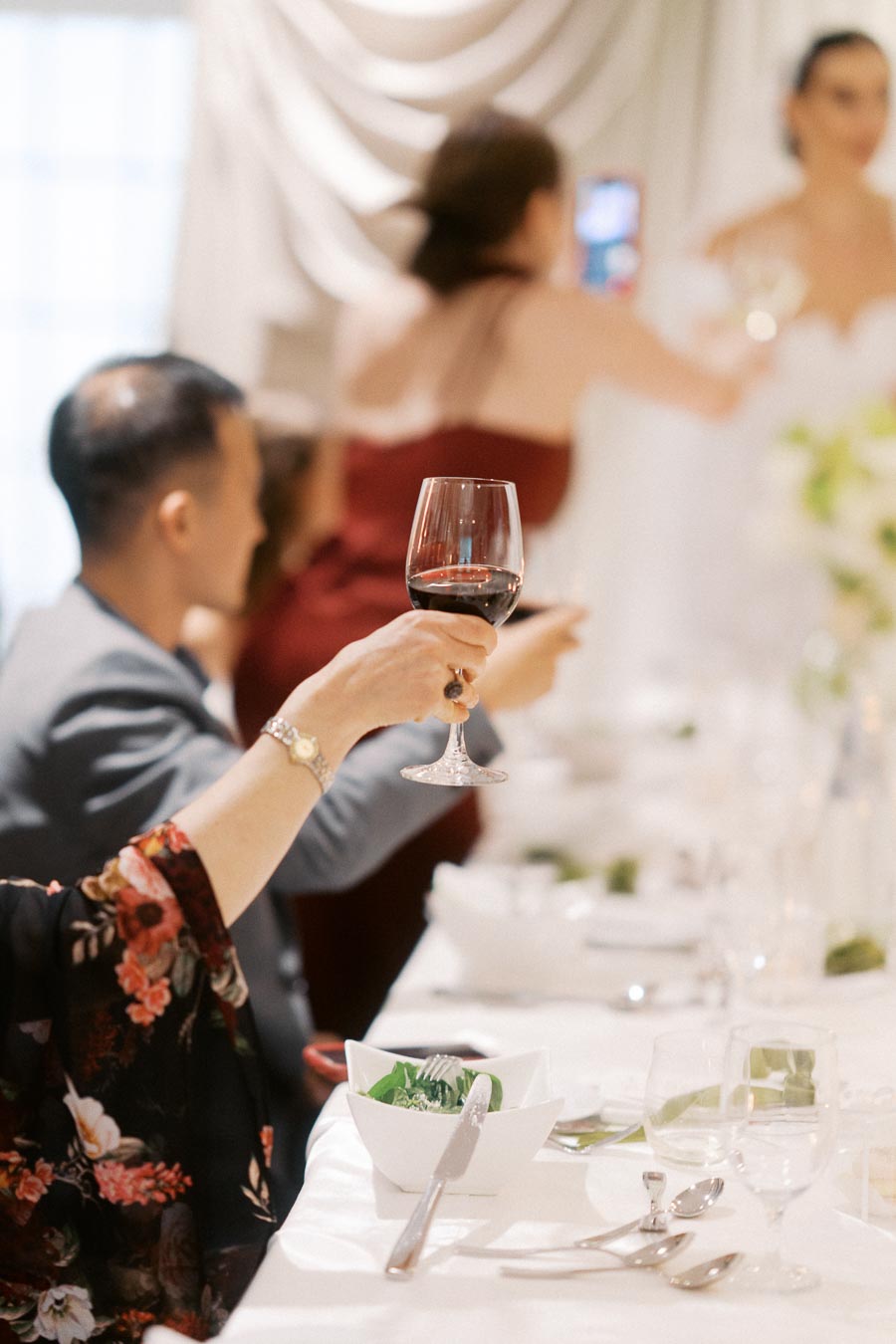 Elegant wedding reception scene with guests raising wine glasses in a toast, featuring a beautifully decorated table and guests in formal attire.