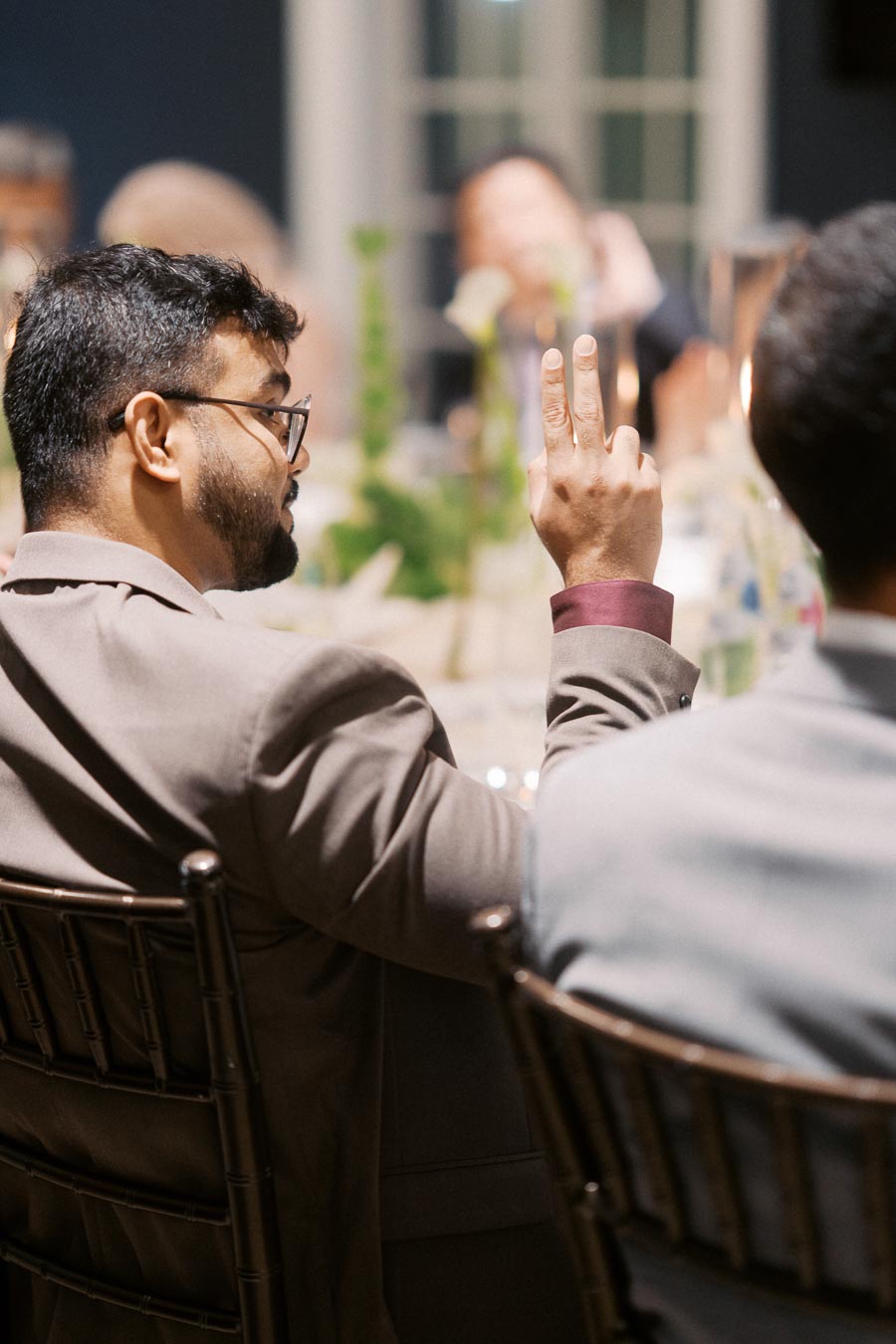 A man in a suit raising two fingers during a formal meeting or dinner, seated at a table with blurred figures in the background.