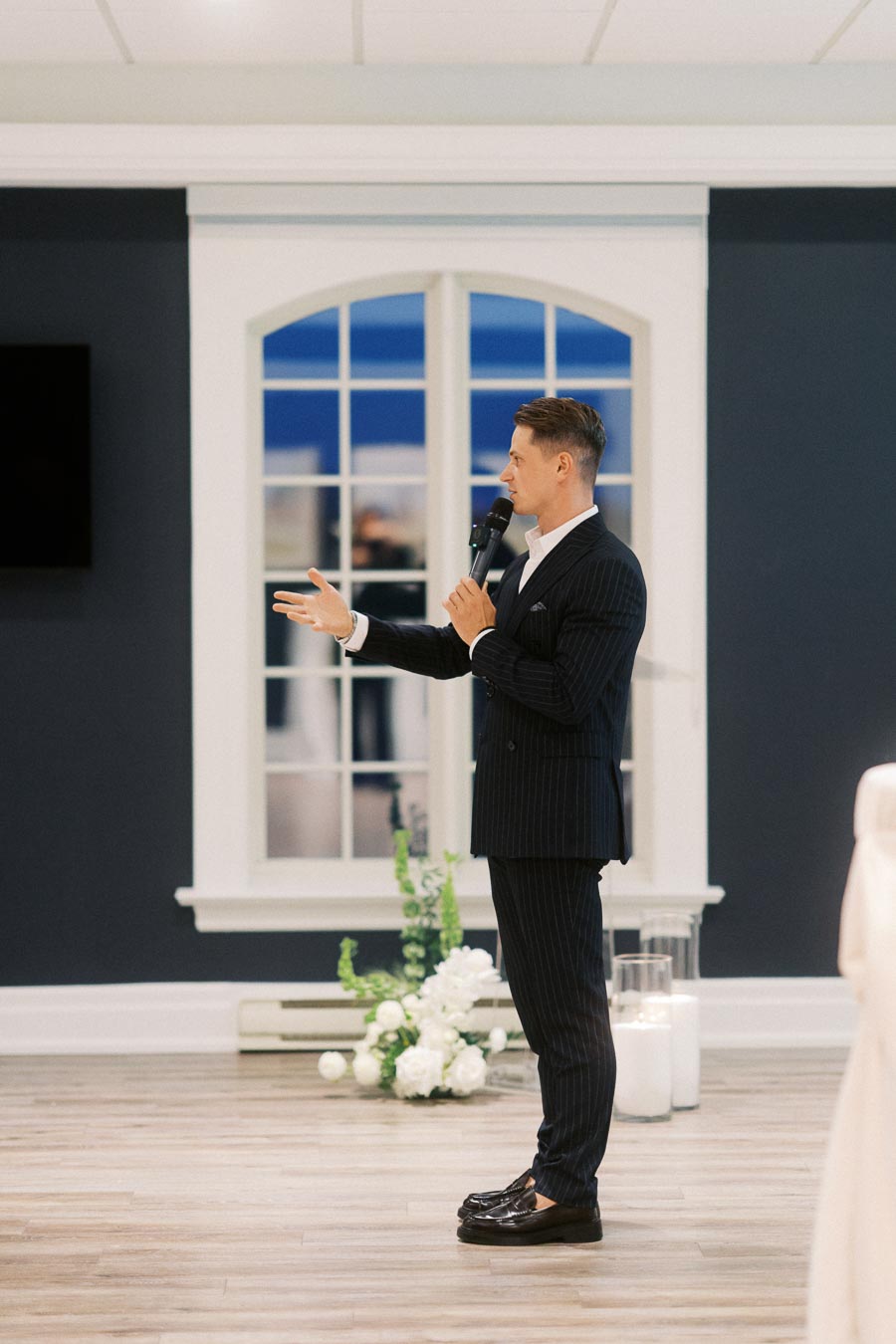 Man in a formal suit speaking into a microphone, standing on a wooden floor in front of a large window, with candles and white flowers in the background.