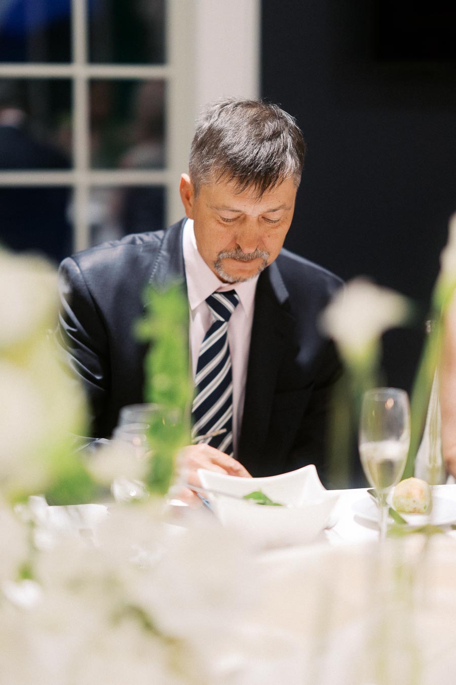 Man in formal attire dining at an event, seated at a table elegantly set with white flowers, a glass of champagne, and a small bread roll.