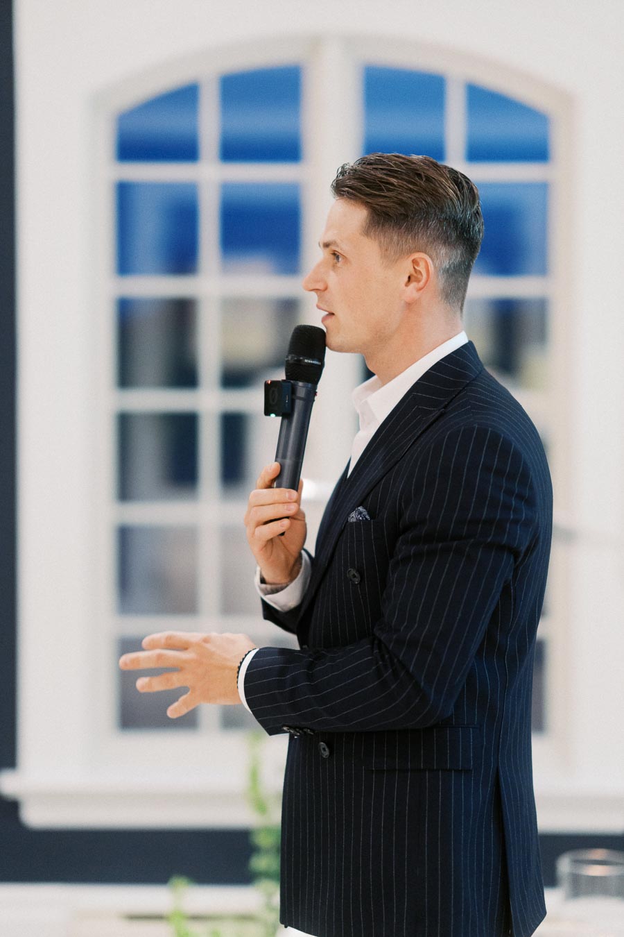Man in a pinstripe suit speaking into a microphone in a bright room with large windows.