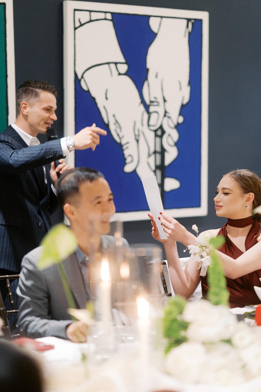 A speaker addresses guests at an elegant dinner event, with contemporary artwork in the background. A woman in a red dress reads a card, while a man sits nearby, surrounded by flickering candles and floral arrangements.