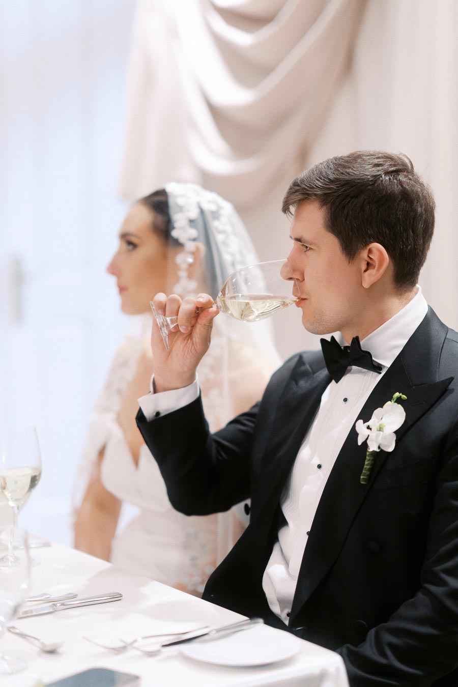 Bride and groom at wedding reception; groom in black tuxedo with orchid boutonniere drinking white wine, bride in white dress and veil, seated at elegantly set table.
