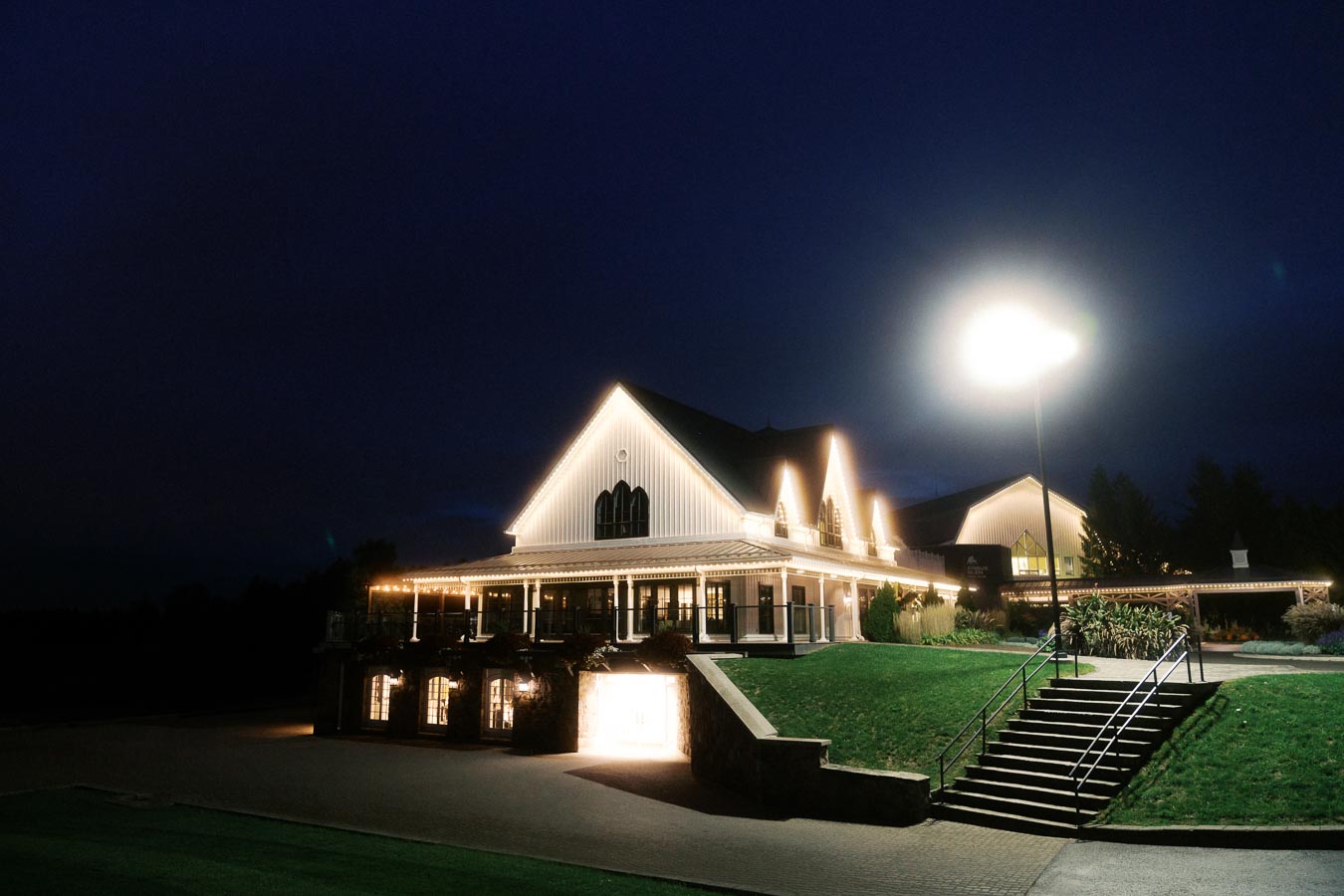 Elegant white building illuminated at night, featuring a lit staircase leading to its entrance, surrounded by well-manicured lawns and a large light post casting an ambient glow, ideal for weddings and events.