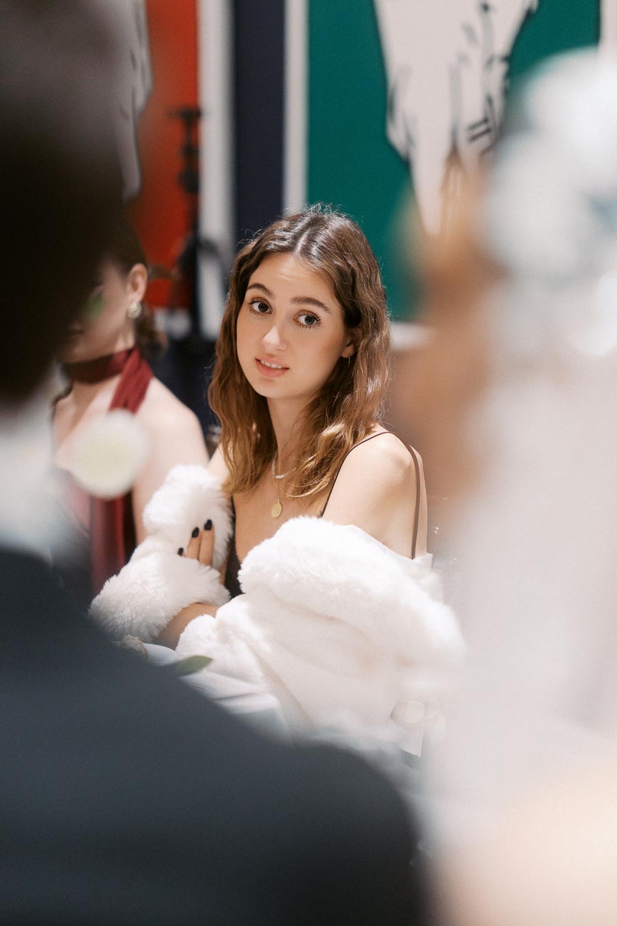 Young woman in elegant attire, wearing a white fur shawl, attending a formal gathering with colorful artwork in the background.
