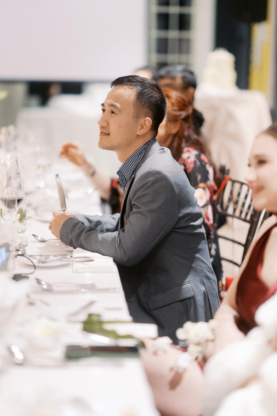 Man in formal attire attentively listens during a dinner event, seated at a elegantly set table with others.
