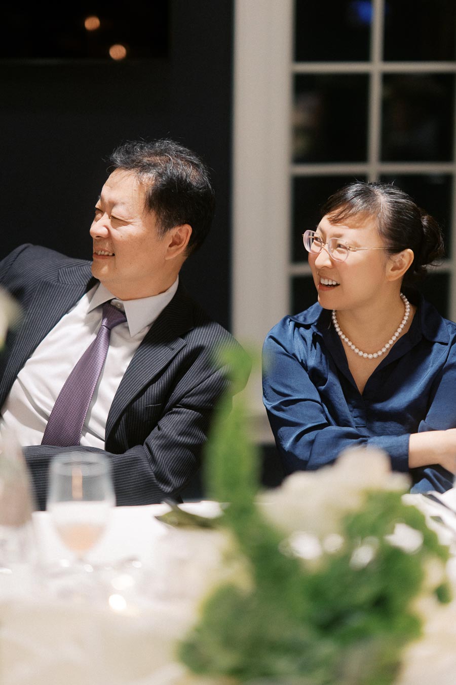 A cheerful couple at a formal event, smiling and sitting at a table with glasses and a floral centerpiece in the foreground.
