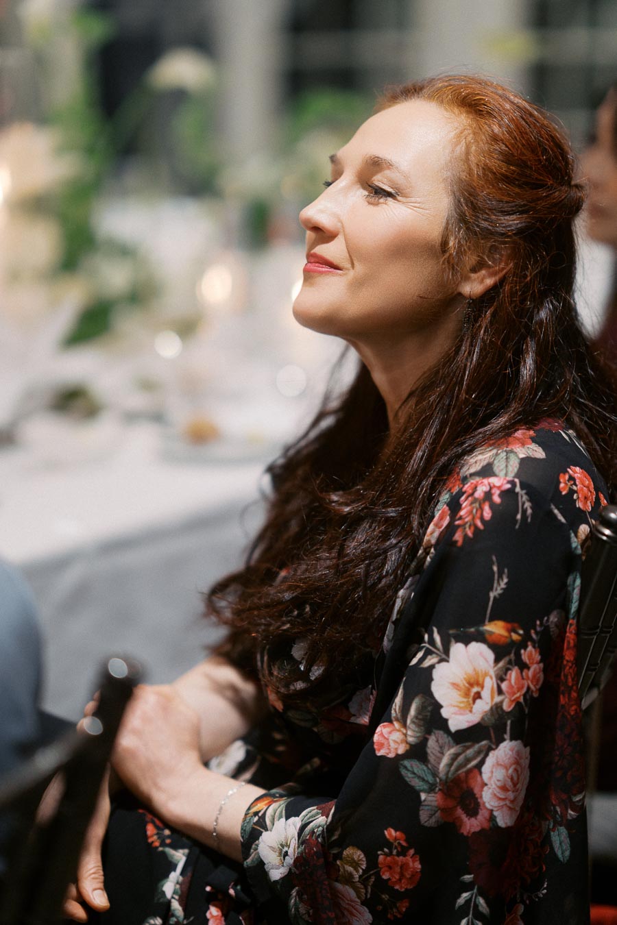 A woman with long red hair in a floral dress sits at a formal event, looking content and relaxed, with a softly blurred background of a decorated table.