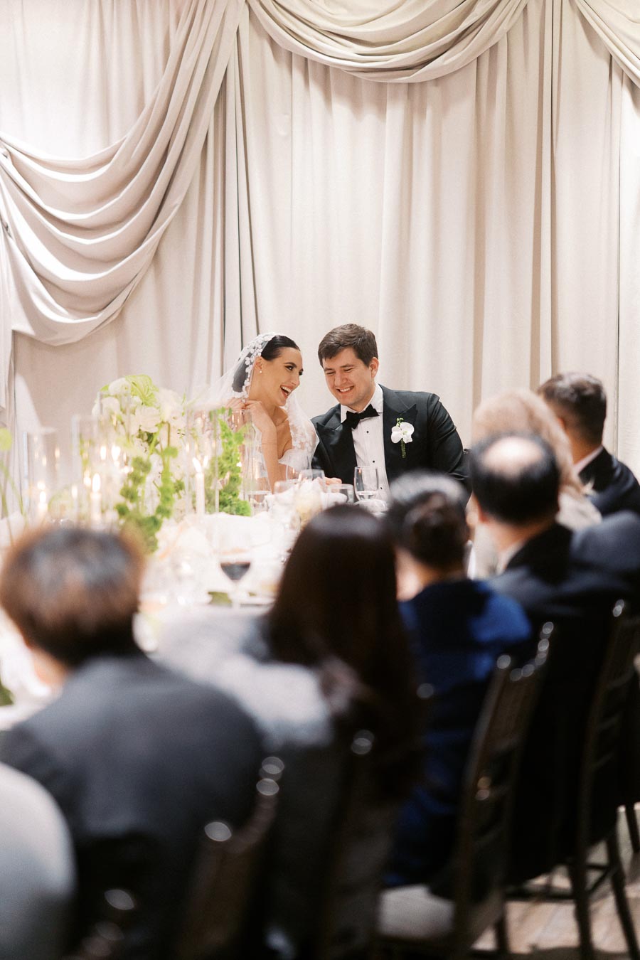 Bride and groom in elegant attire sharing a joyful moment at their wedding reception, surrounded by guests at an elegantly decorated table with draped curtains in the background.