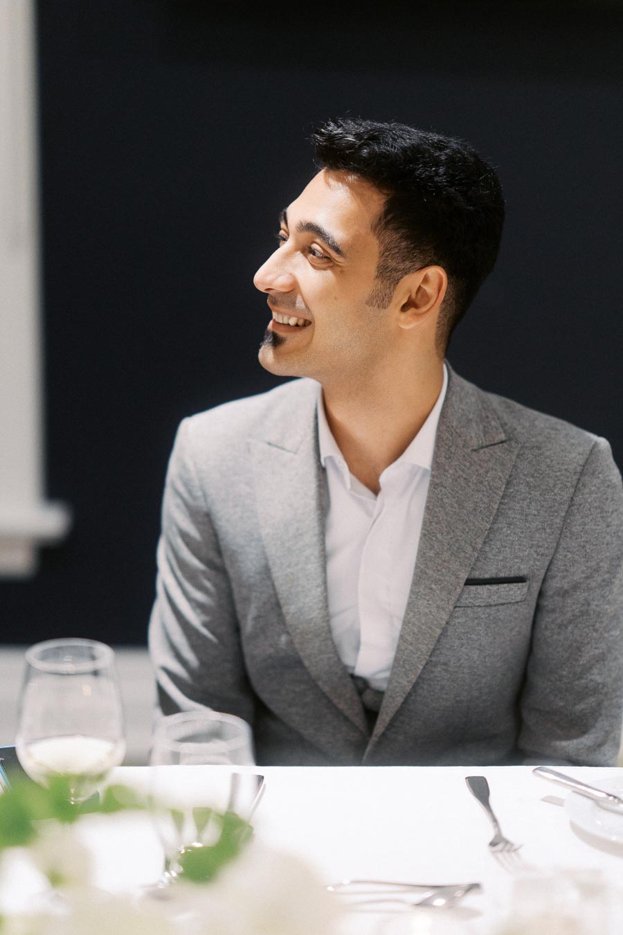 A smiling person in a gray suit sits at a table set with wine glasses and cutlery, looking to the side against a dark background.