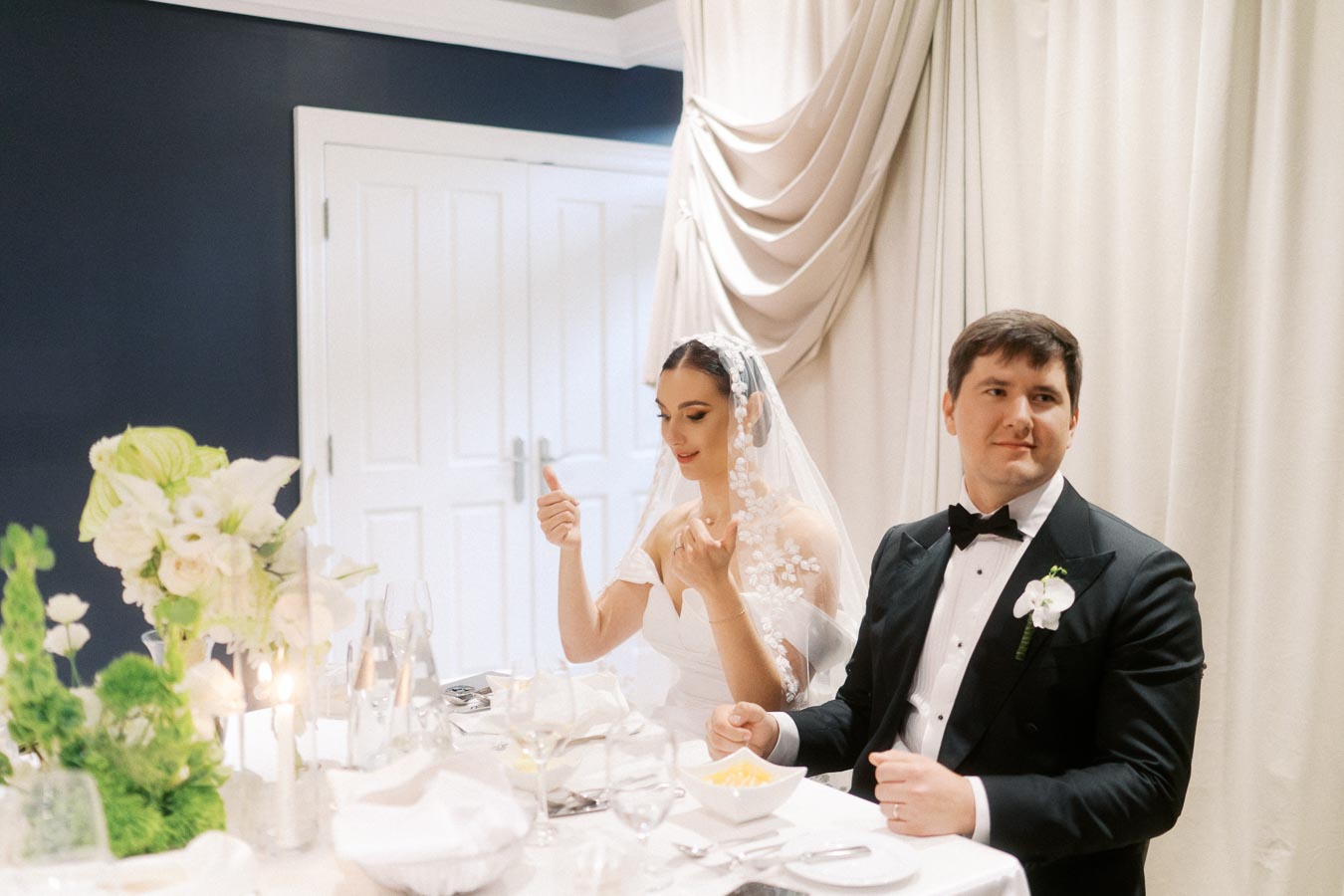 Bride and groom seated at a wedding reception table, adorned with elegant floral arrangements and fine dinnerware, while the bride gestures with a smile and the groom looks on contentedly.