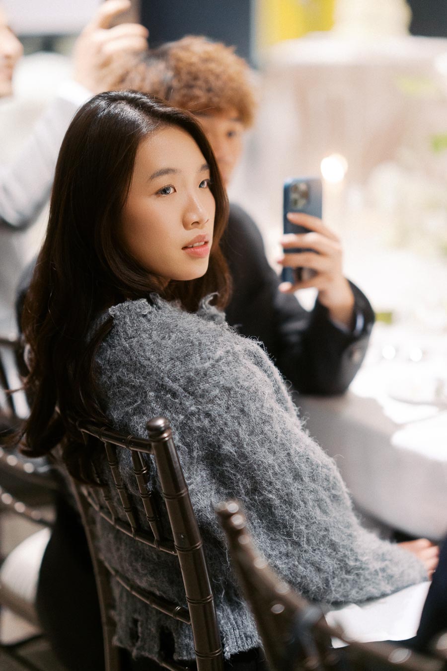 A woman with long dark hair wearing a cozy gray sweater, seated at an event, with a blurred background featuring other attendees and table settings.