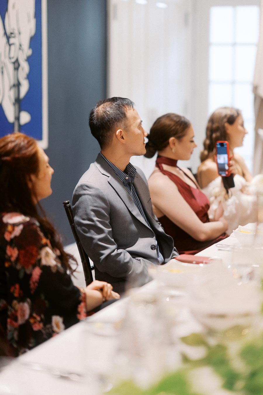 A group of elegantly dressed people seated at a formal dining table, with one person capturing a moment on a smartphone.