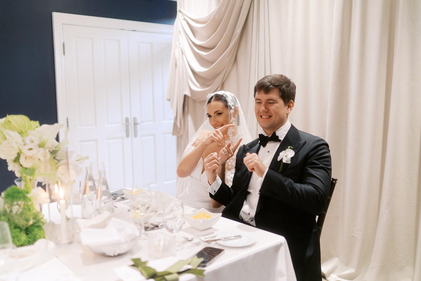 Bride and groom enjoying a lighthearted moment at their wedding reception, sitting at a beautifully decorated table with white floral arrangements and elegant drapery in the background.
