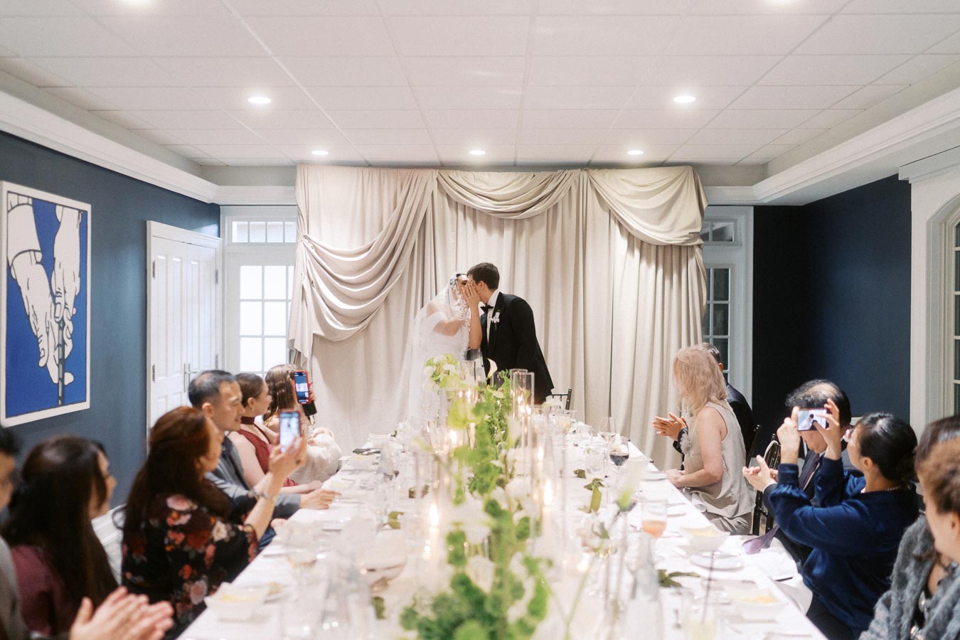 Bride and groom kissing at their wedding reception in a beautifully decorated room, surrounded by guests capturing the moment with their phones, long elegantly set table with greenery centerpiece and candles.