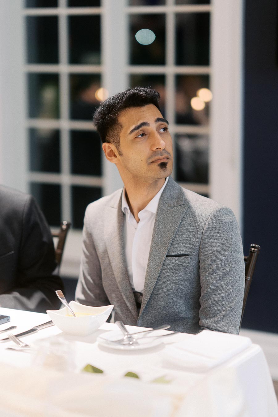 A man in a gray suit sits at a dining table with a thoughtful expression. The background features large windows, creating a formal and elegant ambiance. Ideal for themes of business dining, formal events, or personal reflection.