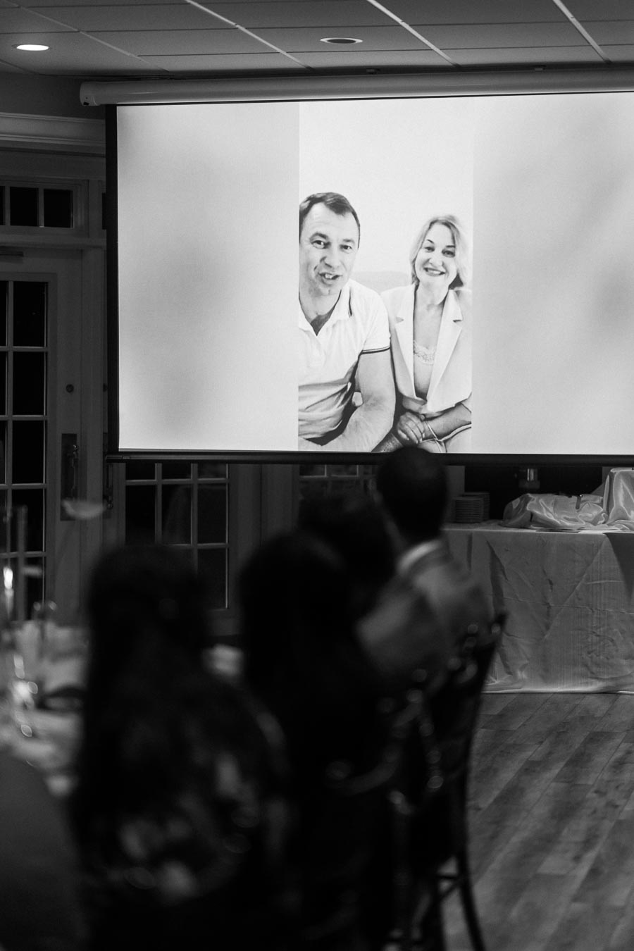 Black and white photo of a large screen displaying a smiling couple in a video call, viewed by an audience in a dimly lit room during an event.