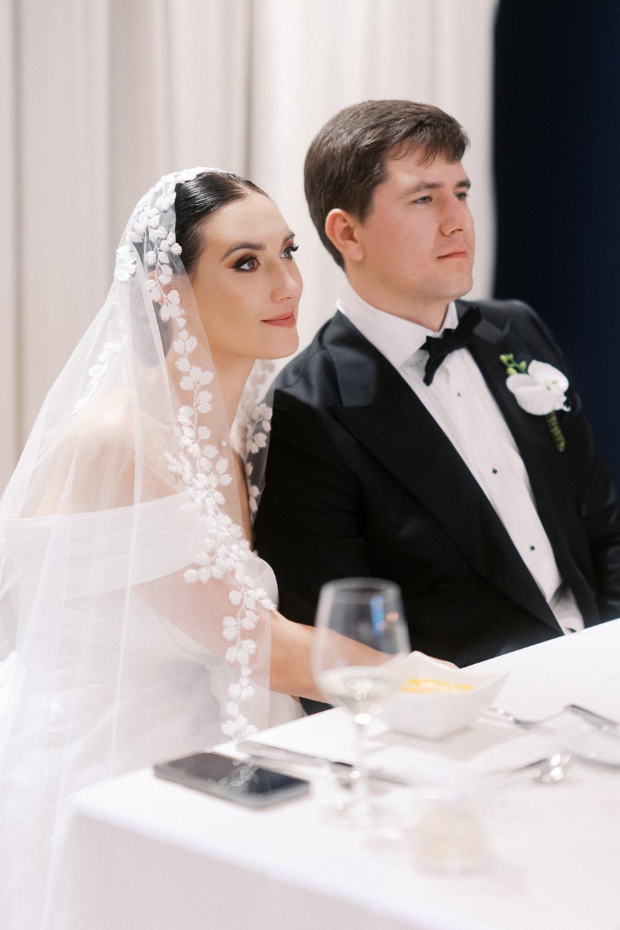 Elegant bride in a floral lace veil sitting beside her groom in a black tuxedo at a wedding reception table.