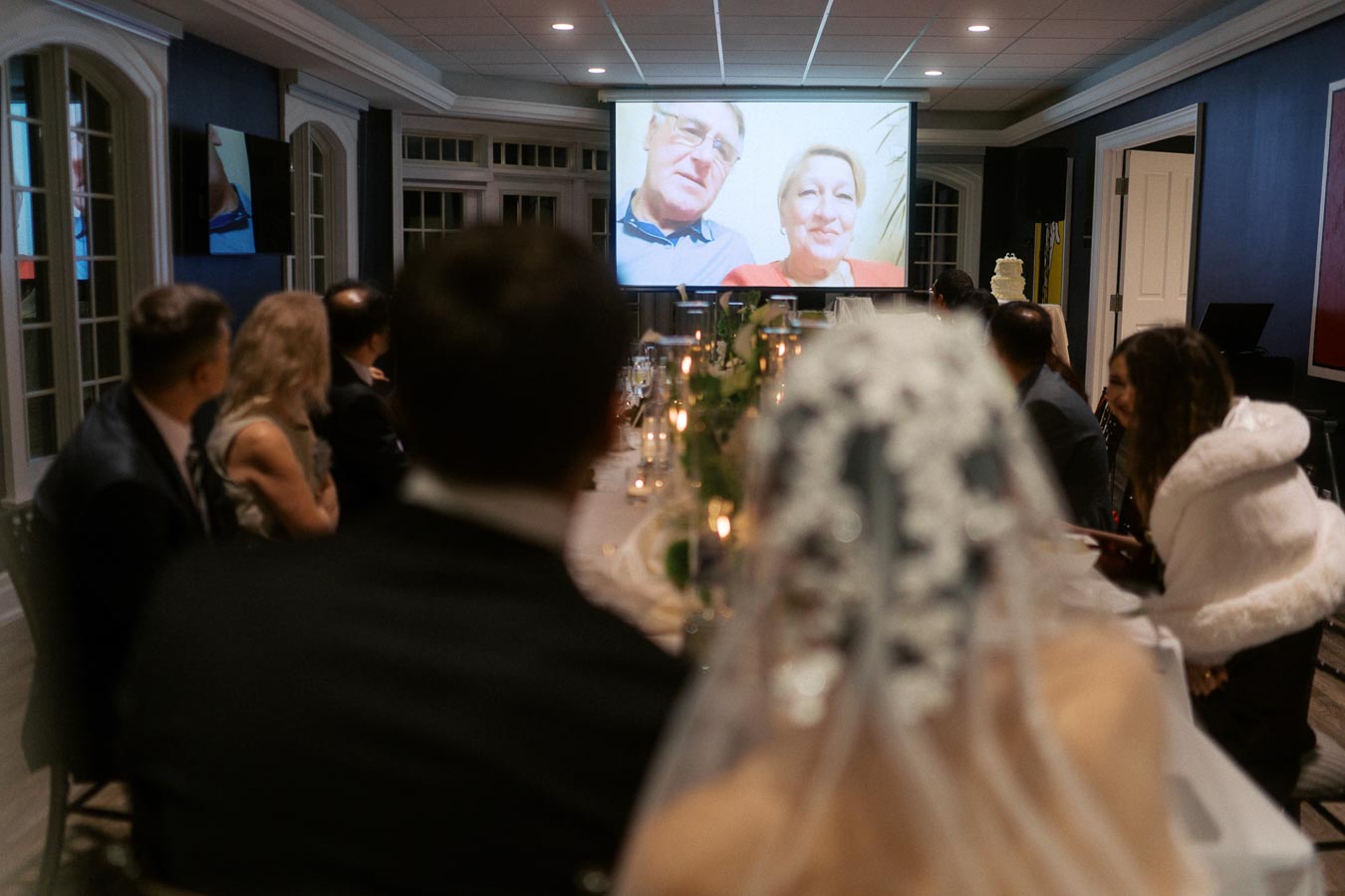Guests watching a wedding video message from family members on a screen during a reception, with a bride and groom seated at a decorated table.