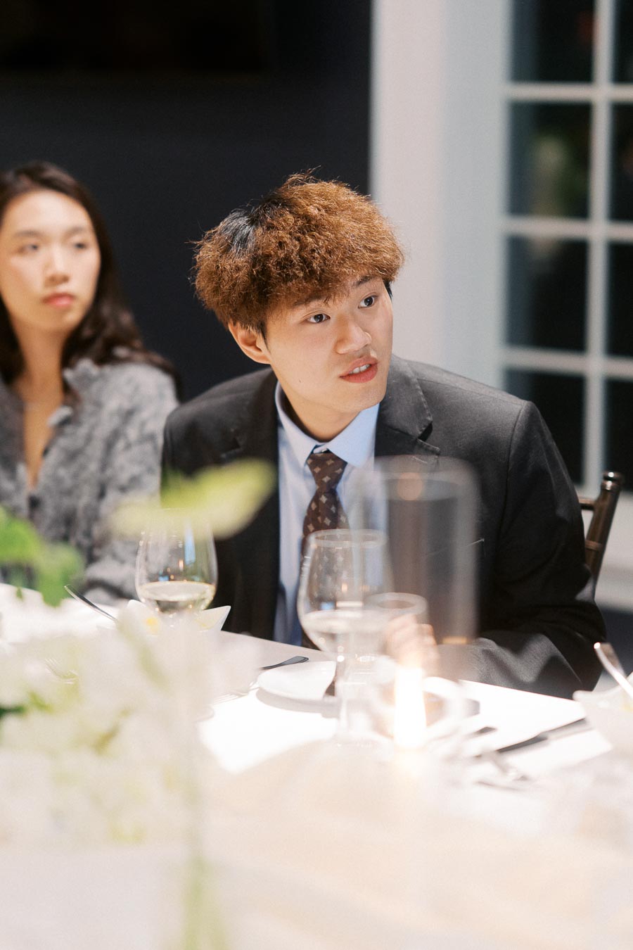 Young man in a formal suit speaking at an elegant dinner event, with a woman sitting beside him, surrounded by a tastefully set table with wine glasses and soft lighting.