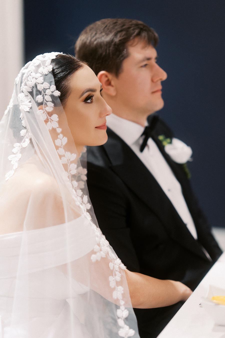 A bride in a white floral lace veil and off-the-shoulder gown sits beside a groom in a black tuxedo, both focused ahead during their wedding ceremony.