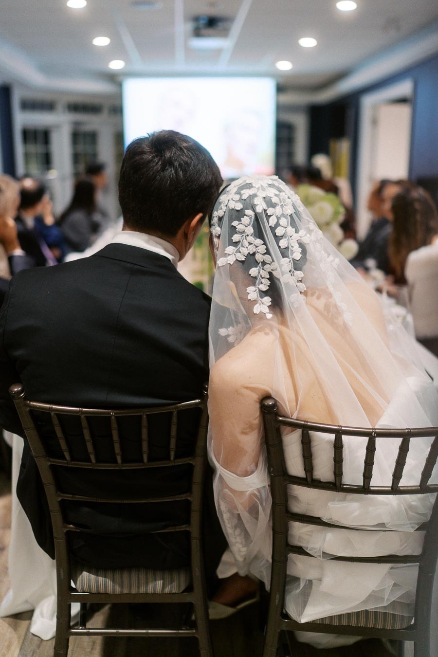 Bride and groom sitting closely at a wedding reception, with the bride wearing a floral lace veil, viewed from behind.