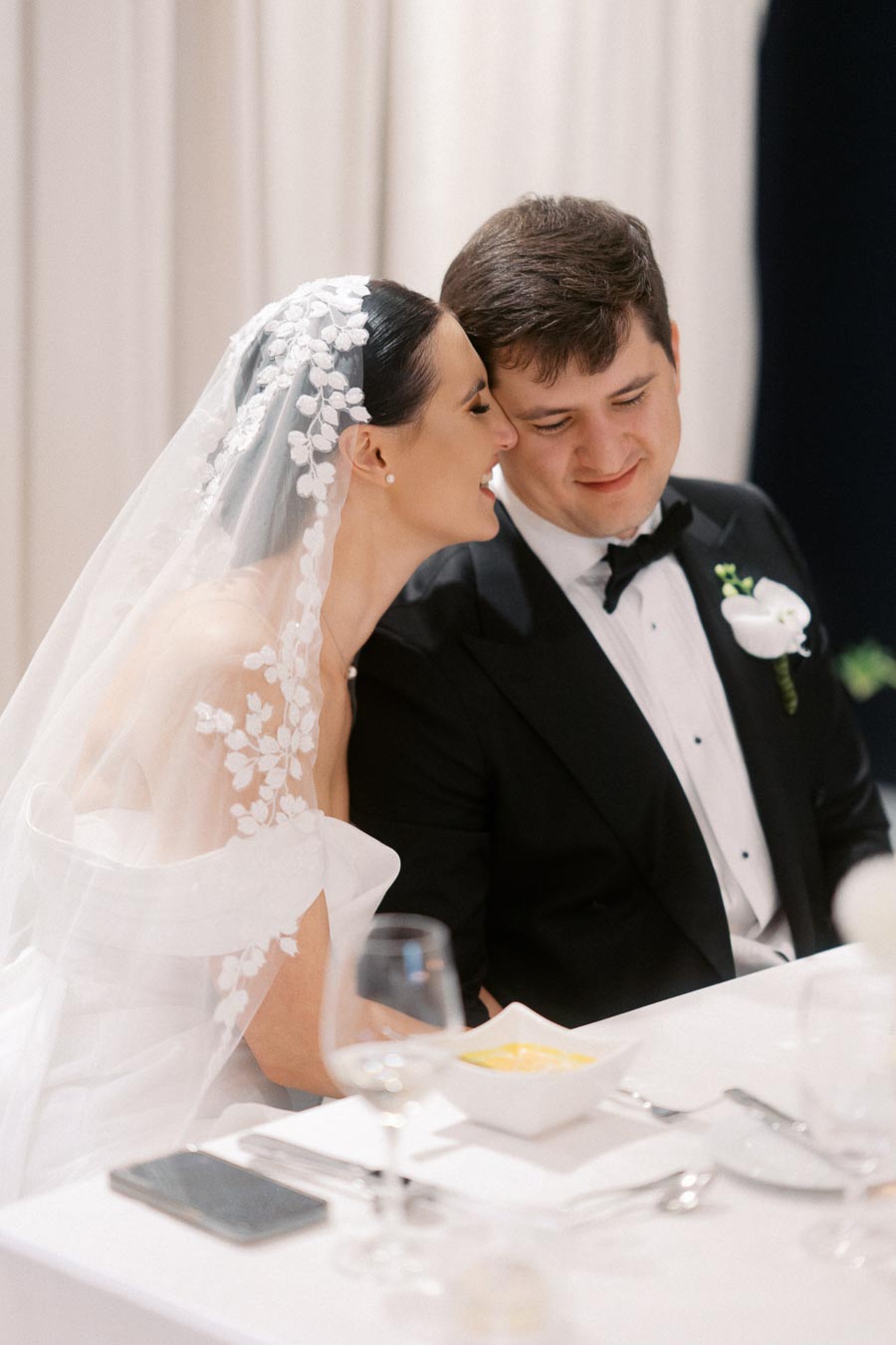 A bride in a white wedding dress with floral veil leans affectionately towards the groom in a black tuxedo during a wedding reception at an elegantly set table.