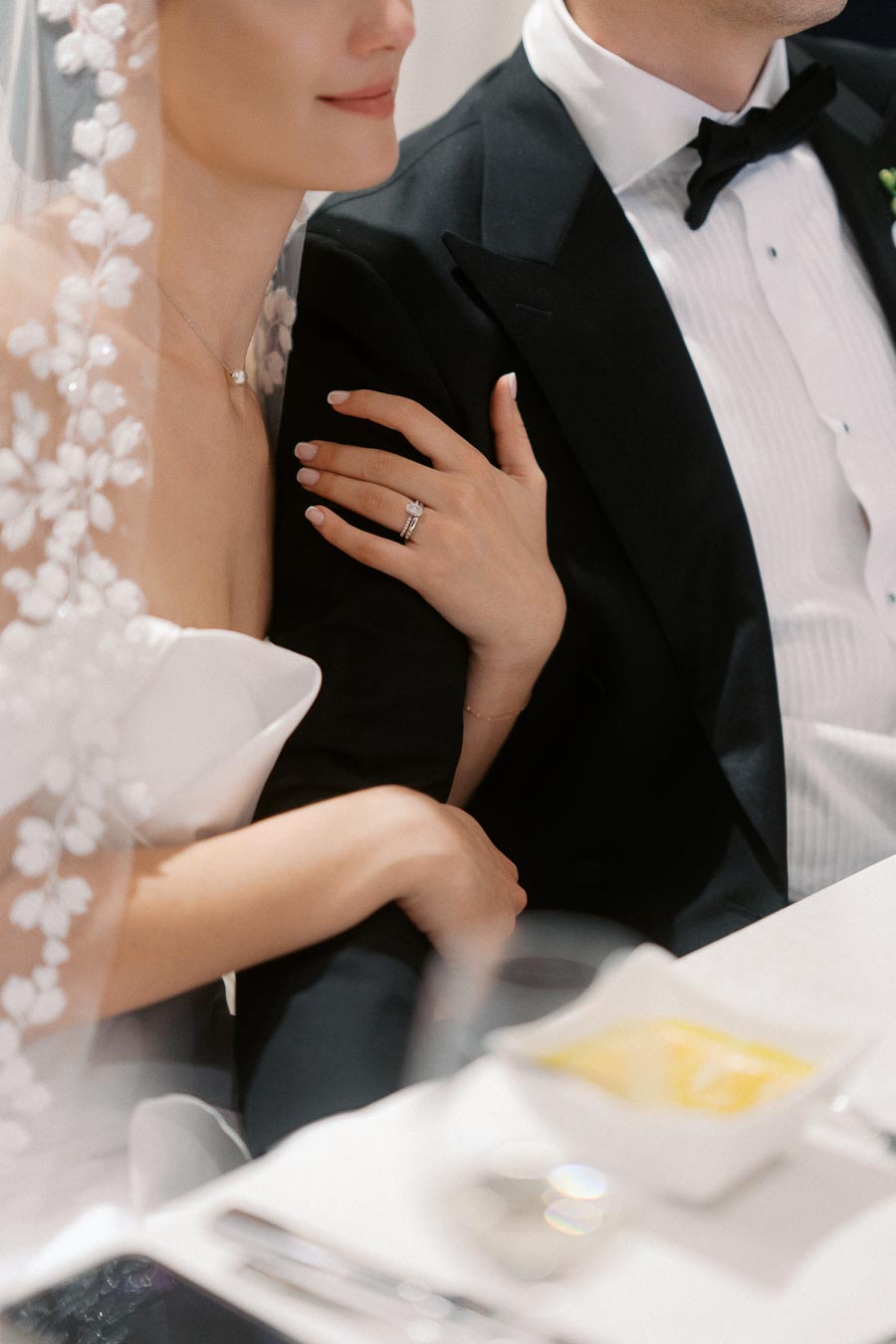 Bride in a floral embroidered veil holding groom's arm during wedding ceremony, showcasing her engagement ring and polished nails.