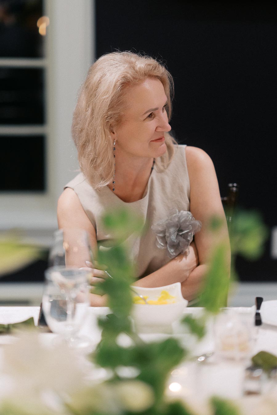 A smiling woman with blonde hair sits at a table during an elegant dinner, wearing a light-colored dress adorned with a decorative flower, surrounded by blurred glassware and green foliage.